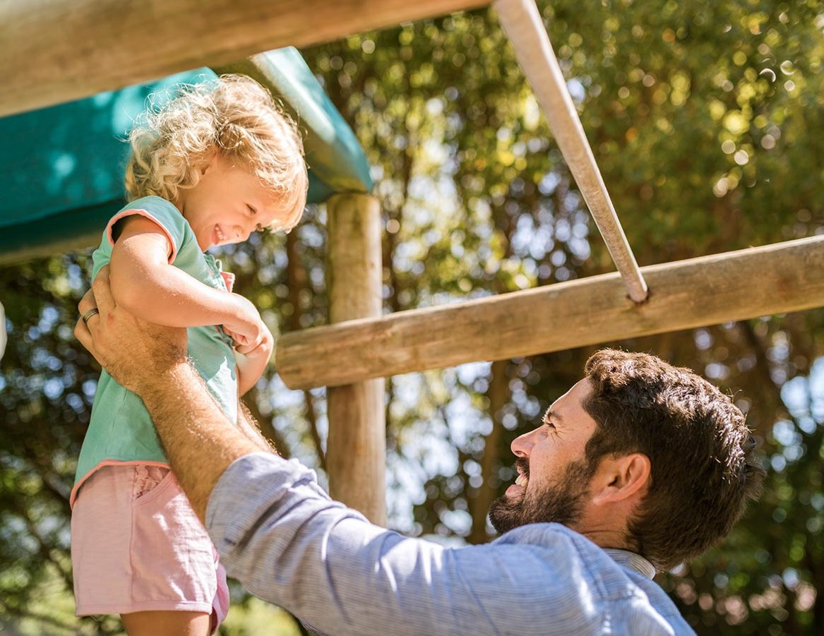 Spielgeräte für die Kleinen gibt es auch bei uns. Vater mit Kind auf Spielplatz.